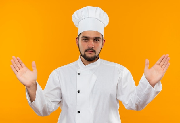 Confident young male cook in chef uniform showing empty hands isolated on orange wall