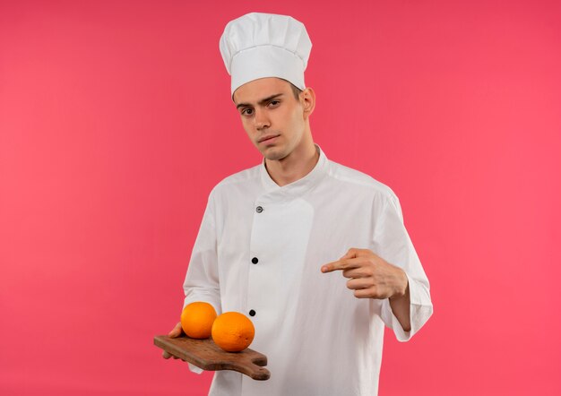 Confident young male cook wearing chef uniform holding and points orange on cotting board on isolated pink wall