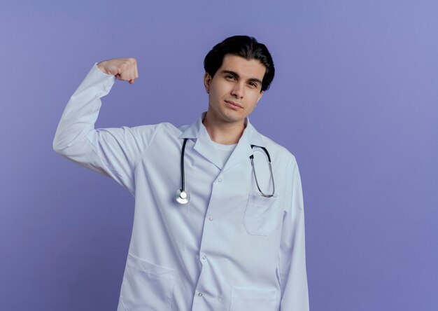 Confident young male doctor wearing medical robe and stethoscope  and doing strong gesture isolated on purple wall with copy space