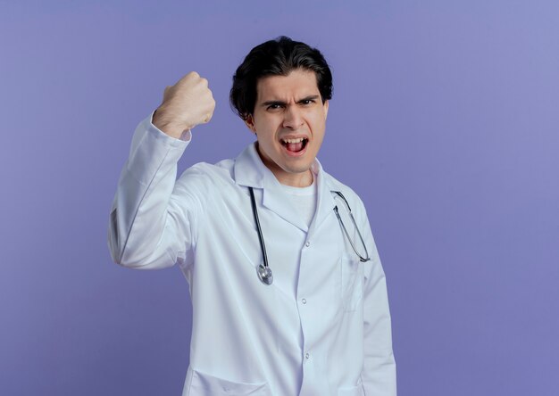 Confident young male doctor wearing medical robe and stethoscope  doing strong gesture isolated on purple wall with copy space