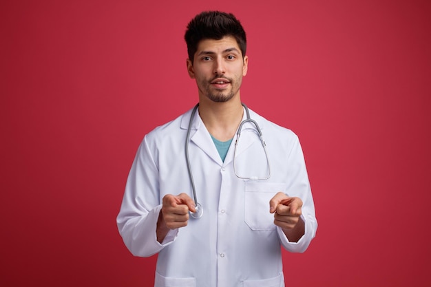 Confident young male doctor wearing medical uniform and stethoscope around his neck looking at camera showing you gesture isolated on red background