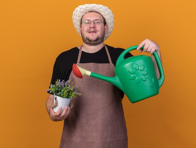 Free Photo confident young male gardener wearing gardening hat and gloves holding watering can with flower in flowerpot isolated on orange wall