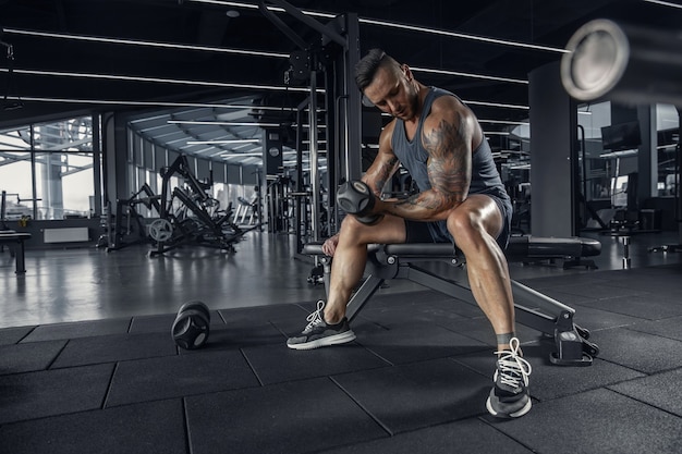 Confident. Young muscular caucasian athlete practicing in gym with the weights. Male model doing strength exercises, training his upper body. Wellness, healthy lifestyle, bodybuilding concept.