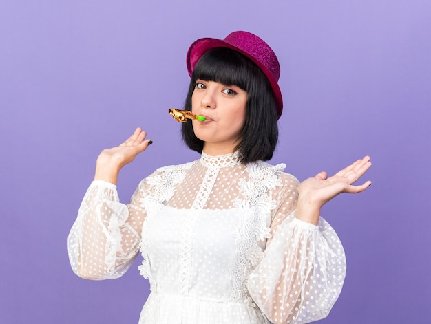 Confident young party girl wearing party hat blowing party horn showing empty hands isolated on purple wall