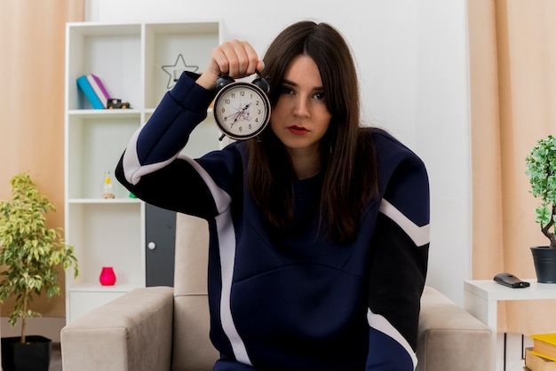 Free photo confident young pretty caucasian woman sitting on armchair in designed living room holding alarm clock near head and looking