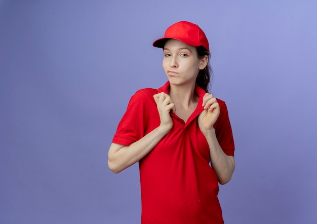 Free photo confident young pretty delivery girl wearing red uniform and cap grabbing collar of her t-shirt isolated on purple background with copy space