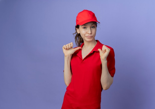 Free photo confident young pretty delivery girl wearing red uniform and cap pointing at herself isolated on purple background with copy space