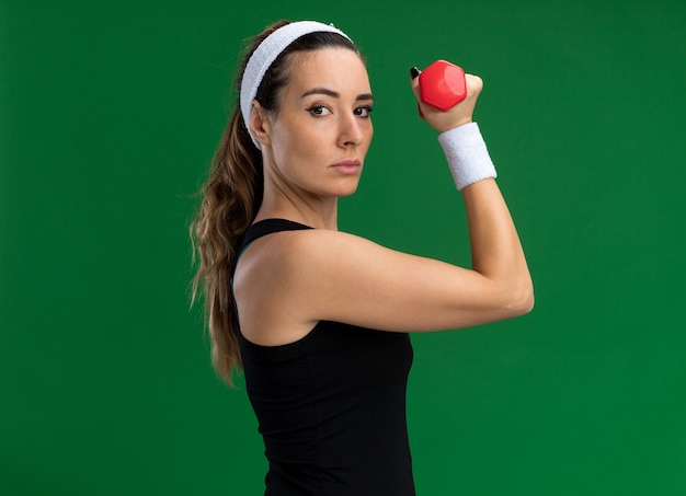 Confident young pretty sporty woman wearing headband and wristbands standing in profile view holding dumbbell