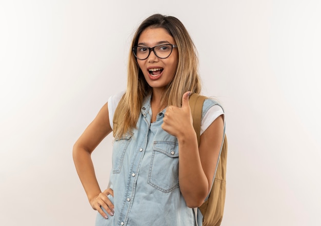 Confident young pretty student girl wearing glasses and back bag putting hand on waist and showing thumb up isolated on white  with copy space