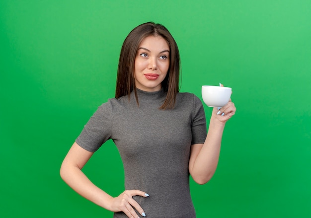 Confident young pretty woman holding cup putting hand on waist looking at side isolated on green background with copy space