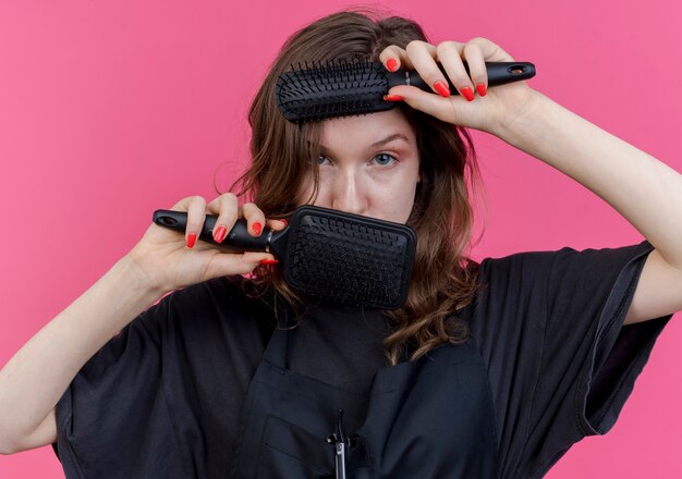 Confident young slavic female barber wearing uniform looking at camera between combs touching face with them isolated on pink background