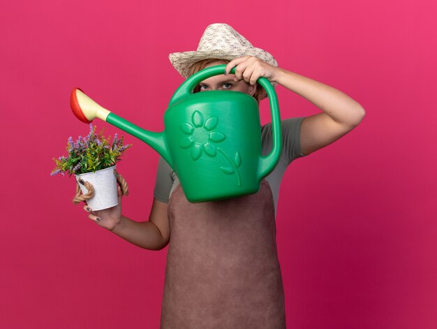 Confident young slavic female gardener wearing gardening hat looking at camera through watering can and holding flowers in flowerpot