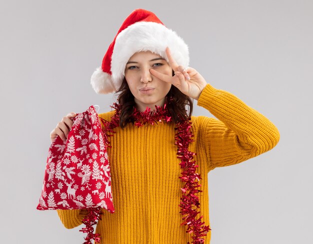 Confident young slavic girl with santa hat and with garland around neck holding christmas gift bag and gesturing victory sign isolated on white background with copy space