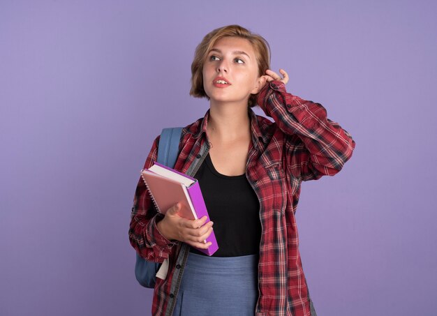 Confident young slavic student girl wearing backpack holds book and notebook looks at side 