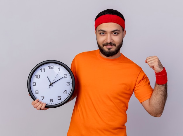 Free photo confident young sporty man wearing headband and wristband holding wall clock showing strong gesture isolated on white background