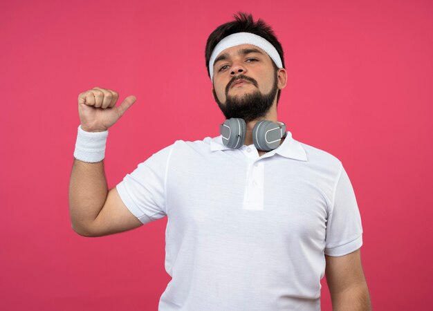 Confident young sporty man wearing headband and wristband with headphones points at himself