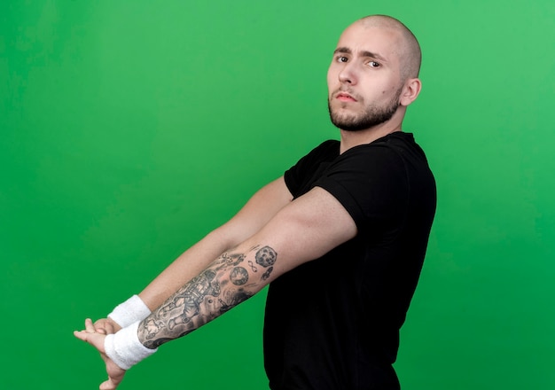 Free photo confident young sporty man wearing wristband stretching out arms isolated on green wall