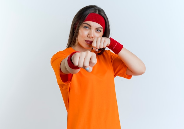 Confident young sporty woman wearing headband and wristbands  doing boxing gesture isolated on white wall with copy space