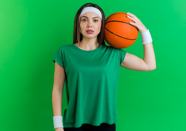 Free photo confident young sporty woman wearing headband and wristbands holding basketball ball on shoulder looking