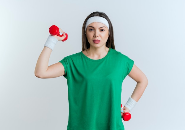 Confident young sporty woman wearing headband and wristbands holding dumbbells looking 