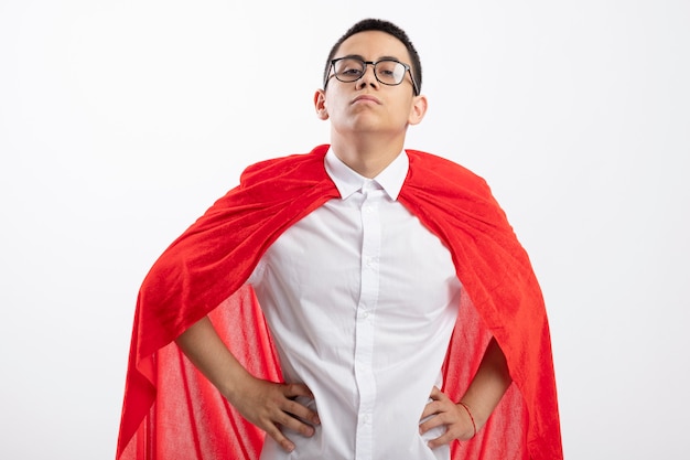 Free photo confident young superhero boy in red cape wearing glasses looking at camera keeping hands on waist isolated on white background