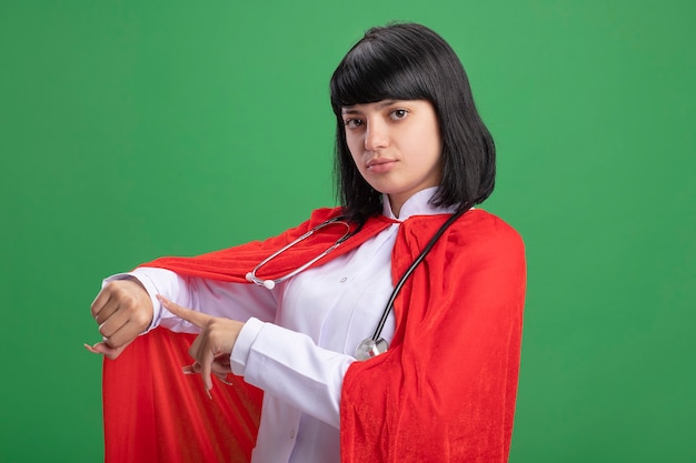 Free photo confident young superhero girl wearing stethoscope with medical robe and cloak showing wrist clock gesture isolated on green wall