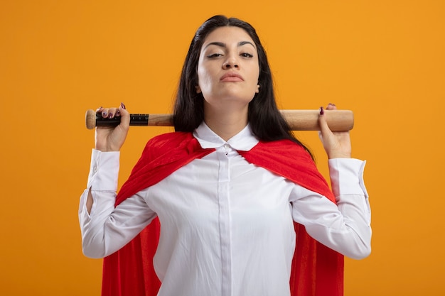 Confident young superwoman holding baseball bat behind neck looking at front isolated on orange wall