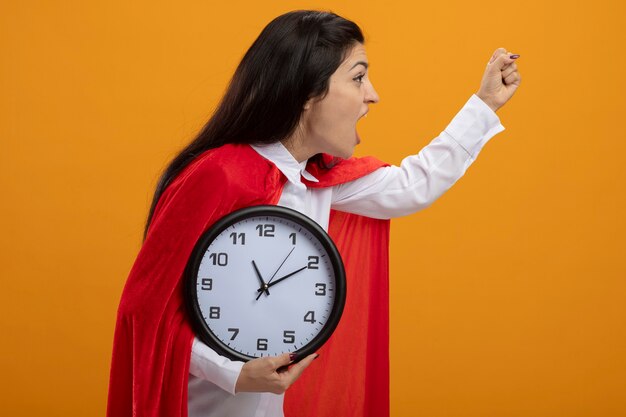 Confident young superwoman standing in profile view holding clock raising fist up looking straight isolated on orange wall