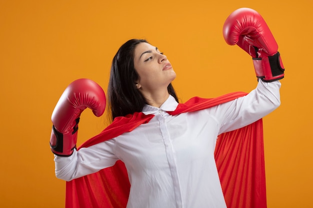 Free photo confident young superwoman wearing box gloves doing strong gesture looking at her hand isolated on orange wall