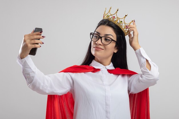 Confident young superwoman wearing glasses and crown touching crown taking selfie isolated on white wall