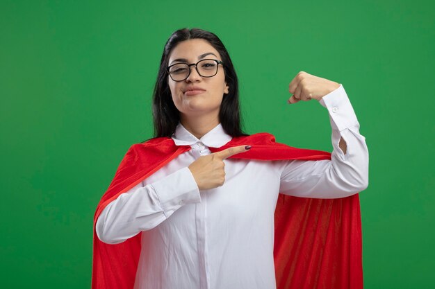 Confident young superwoman wearing glasses flexing her muscles and getting fun looking at front isolated on green wall