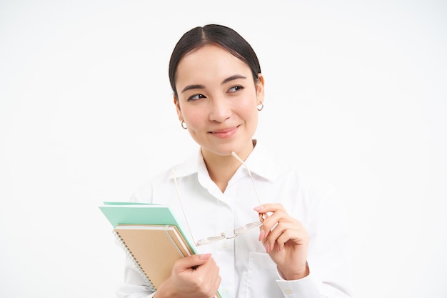 Free photo confident young woman office worker with glasses holds notebooks looks thoughtful thinking standing