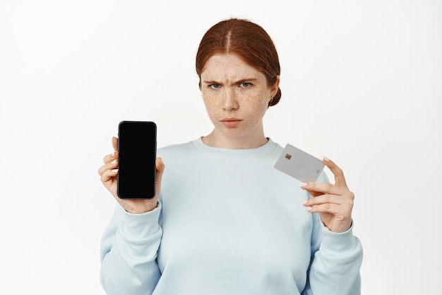 Confused or angry redhead girl showing credit card with mobile phone screen on white