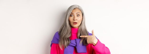 Free photo confused asian senior woman with grey hair gasping startled pointing at herself and looking at camer