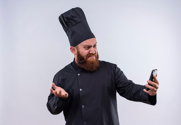 A confused bearded chef man in black uniform showing angry expression while looking at his mobile phone on a white wall