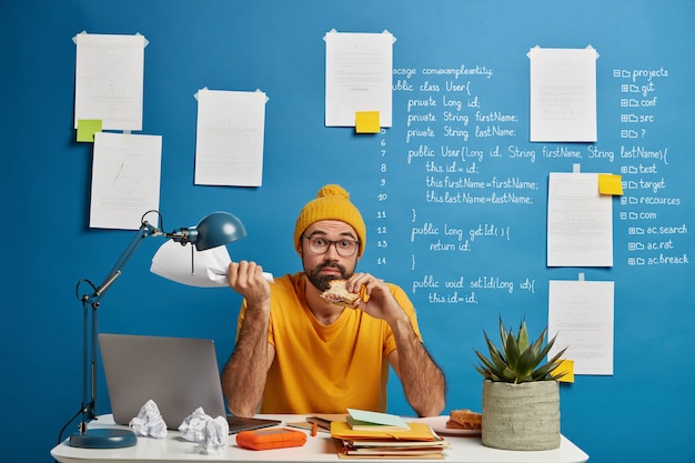 Free Photo confused bearded freelance worker develops startup project, has lunch, eats delicious snack, holds paper document in hand, poses at desktop.