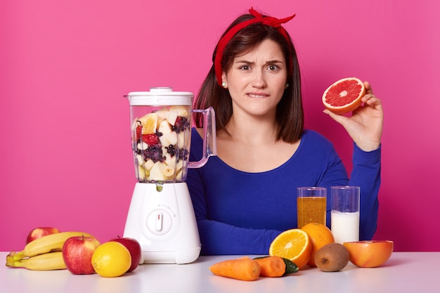 Confused black haired emotional female sits at table, biting her lip, holding half of grapefruit in one hand, mixing fruit in blender, making sweet nutritious smoothie for healthy meal on diet.