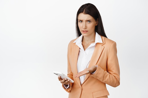 Confused businesswoman pointing at her cellphone staring puzzled at camera standing in suit over white background