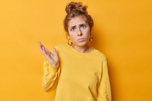 Confused doubtful young European woman with combed hair raises palm and looks puzzled reacts on something unexpected wears casual jumper poses against vivid yellow background So what to do now