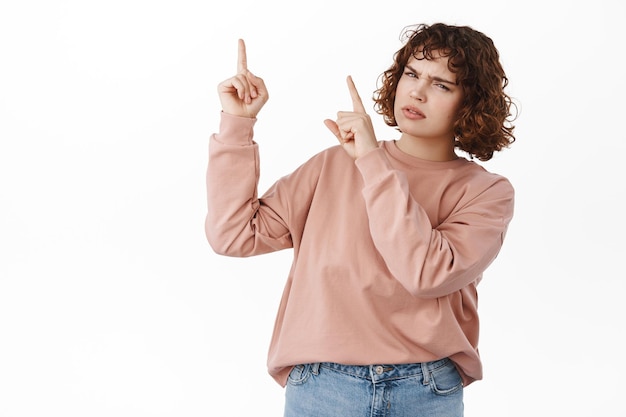 Free photo confused girl points at upper left corner, looks puzzled and clueless, need answers, asking question about advertisement on top, what is this, standing over white background