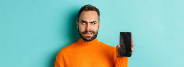 Confused guy scartching head and showing smartphone screen looking puzzled standing over turquoise b