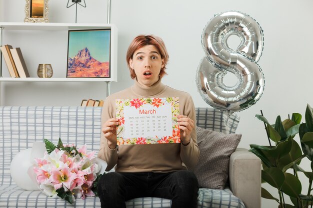 Confused handsome guy on happy women day holding calendar sitting on sofa in living room