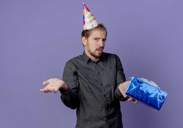 Confused handsome man in birthday cap holds gift box isolated on purple wall