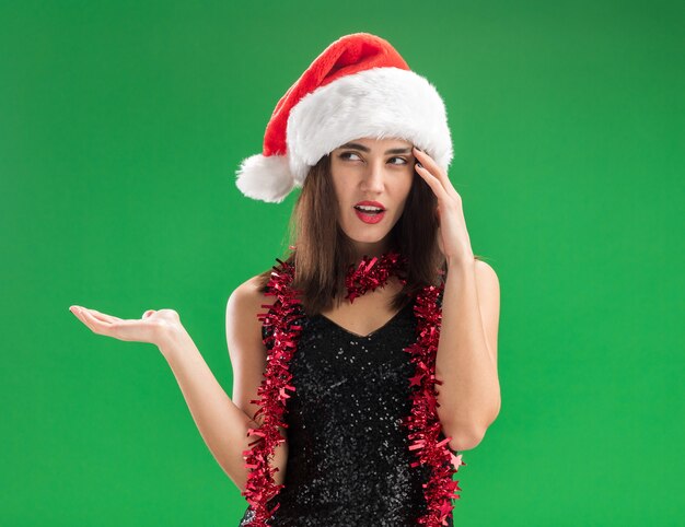 Confused looking side young beautiful girl wearing christmas hat with garland on neck spreading hand isolated on green wall