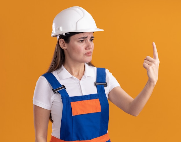 Free Photo confused looking at side young builder woman in uniform points at up isolated on orange wall