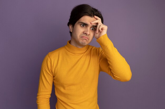 Confused looking at side young handsome guy wearing yellow turtleneck sweater putting hand on temple isolated on purple wall