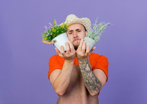 Confused male gardener wearing gardening hat holds flowers in flowerpots 