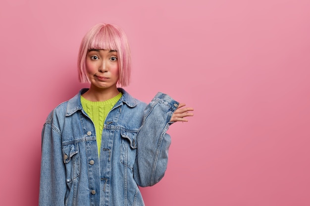 Free photo confused pink haired woman raises hand and stands unaware, has stylish hairstyle, dressed in denim clothes, has no idea, faces dilemma, poses