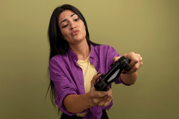 Confused pretty brunette woman holds and looks at game controller isolated on olive green wall