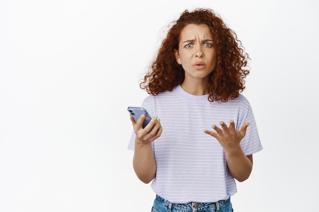 Free Photo confused redhead girl looking puzzled, holding smartphone, cant understand smth on phone, standing over white background